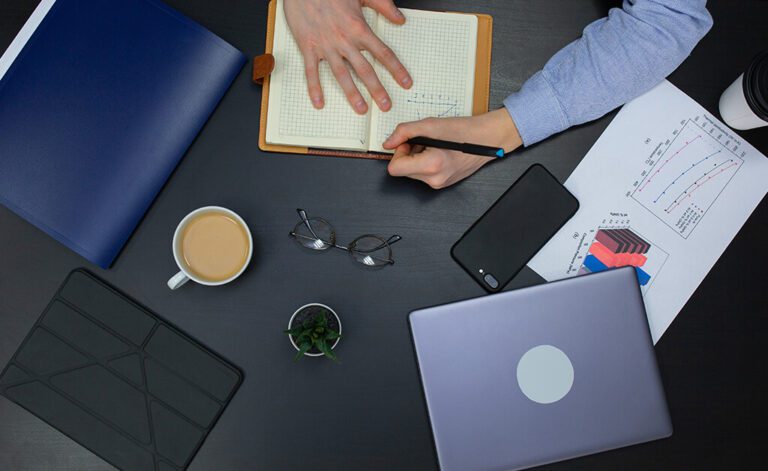 Overhead shot of desk with a laptop and a man's hands writing in a notebook and examining business reporting and analytics.